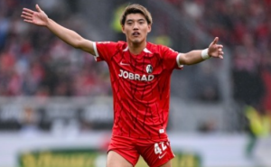 SCフライブルクの赤いユニフォームを着て、試合中に両手を広げて味方に指示を出す（または感情をあらわにする）堂安律