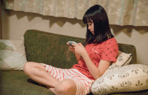 映画「幼な子われらに生まれ」のワンシーン。赤いTシャツにボーダー柄のショートパンツという部屋着姿で緑色のソファに座り、うつむき加減でスマートフォンを操作している南沙良さん。
