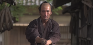 映画『侍タイムスリッパー』の劇中シーン。幕末の侍・高坂新左衛門(山口馬木也)が、まげを結った姿で真剣な面持ちで刀を構え、今まさに斬りかかろうとする緊迫した場面。