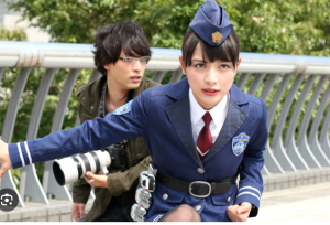 『仮面ライダードライブ』にて、凛とした警察官の制服姿で前を見据える内田理央。背後にはカメラを構えた男性の姿があり、緊迫した現場の空気感と彼女の強い意志が伝わる一枚。