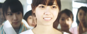 「乃木坂46時代の西野七瀬。白の衣装を身にまとい、仲間たちに囲まれながら、儚げながらも決意を秘めた表情で前を見つめるアップのカット」
