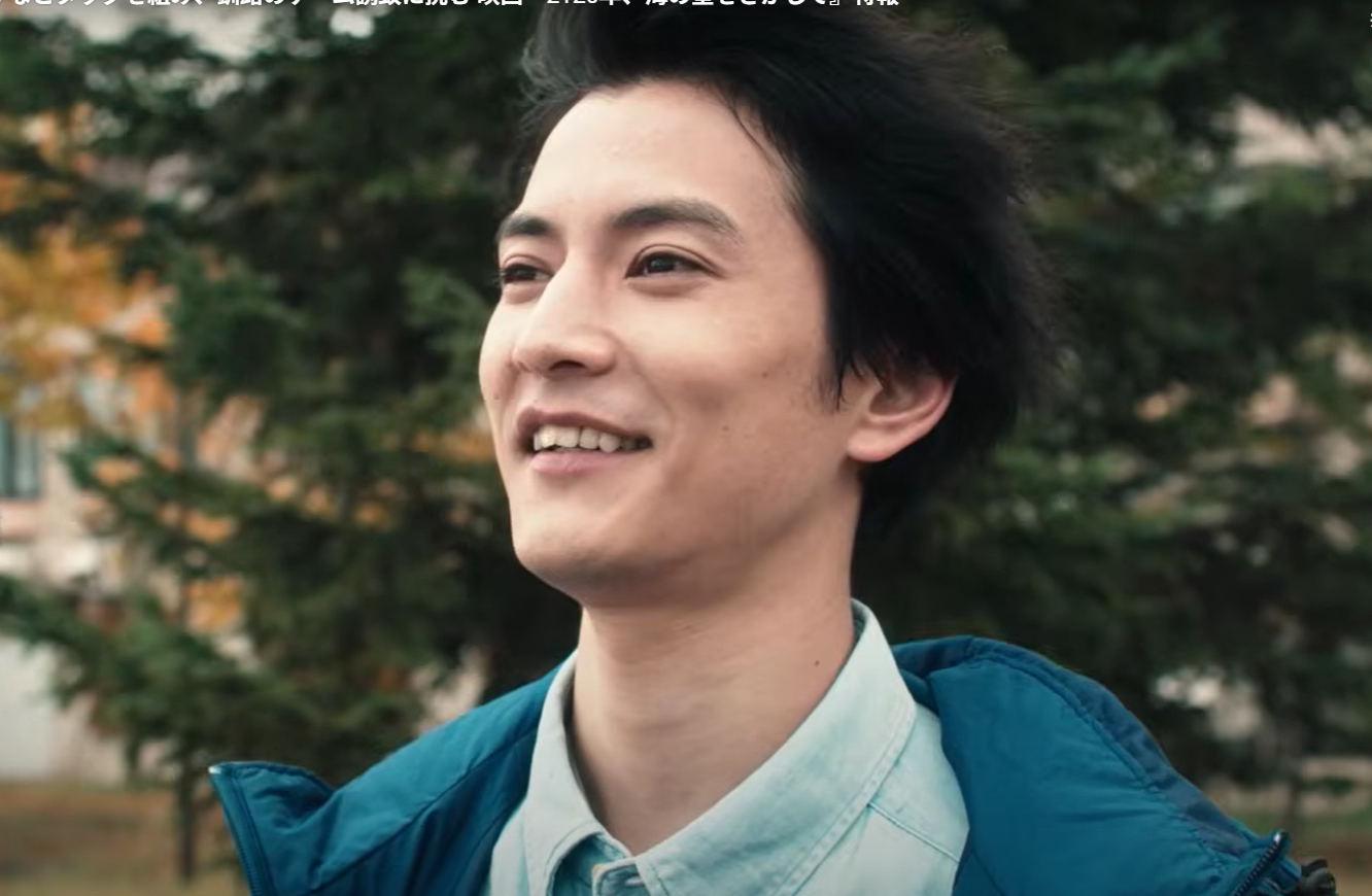 屋外で爽やかな笑顔を見せる俳優の渡邊圭祐。青いダウンジャケットとライトブルーのシャツを着用し、自然光の中で斜め上を見上げている。