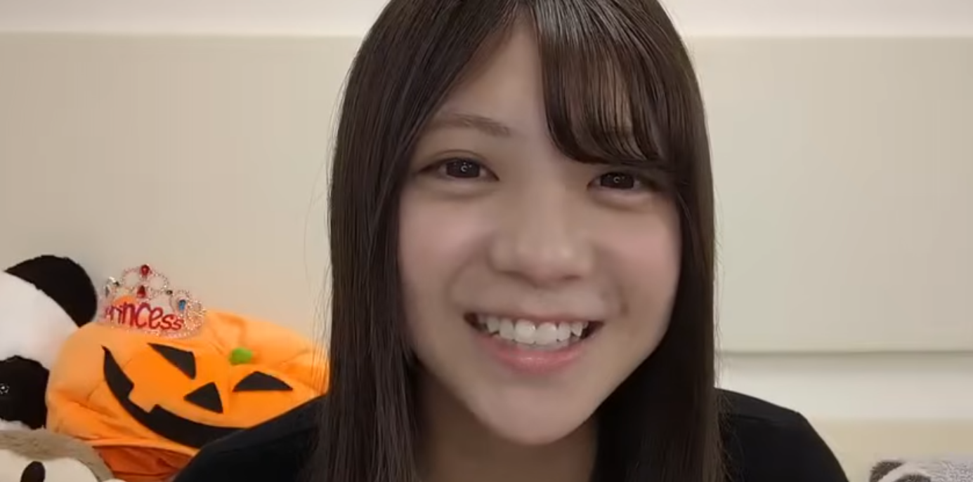 カメラに向かって満面の笑みを浮かべる乃木坂46の5期生・冨里奈央。背景にはハロウィンのカボチャのぬいぐるみやティアラが置かれている。