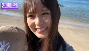 YouTubeチャンネル「乃木坂配信中」のロケにて、砂浜を背景にカメラを覗き込み、弾けるような満面の笑みを見せる小川彩。活動を楽しみ、ファンを魅了する彼女の素の表情が捉えられた至近距離のポートレート。