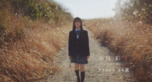 乃木坂46加入直後、千葉県の自然豊かな道を制服姿で歩く14歳の小川彩。「千葉県出身 14歳」という紹介テロップと共に、トップアイドルへの第一歩を踏み出した少女の決意を感じさせるドキュメンタリー映像のワンシーン。