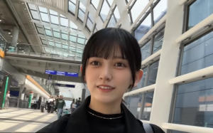 近代的な空港または駅の構内で自撮りをする乃木坂46の池田瑛紗。黒いハイネックのトップスにネックレスを着用し、カメラを見て微笑んでいる。背景には特徴的なアーチ型のガラス屋根と、歩く人々の姿が写っている。