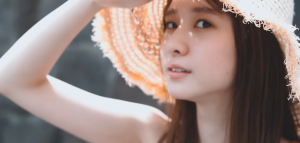 麦わら帽子を被り、右手で帽子のつばを押さえながら、強い日差しの下でこちらを真っ直ぐに見つめる菅原咲月。透明感のある表情が印象的なカット。