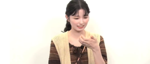 ベージュのベストを着用した乃木坂46の五百城茉央が、右手を口元に寄せ、何かを美味しそうに食べている最中に微笑む瞬間。彼女のおっとりとした親しみやすいキャラクターと日常の幸せを伝えるオフショット。