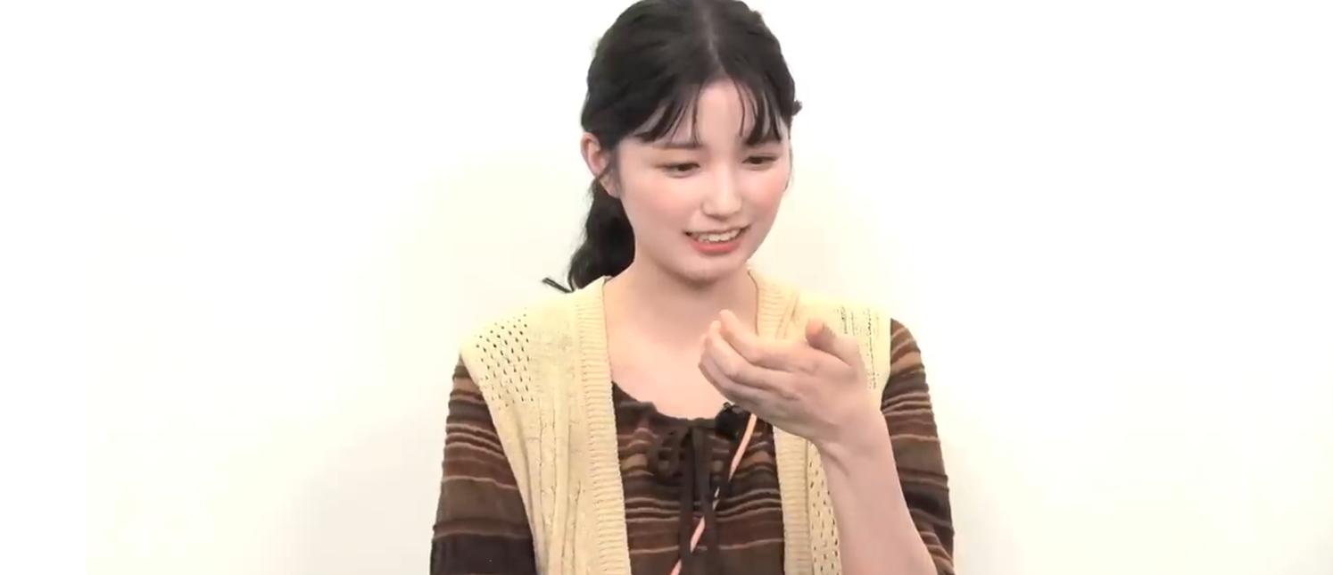 ベージュのベストを着用した乃木坂46の五百城茉央が、右手を口元に寄せ、何かを美味しそうに食べている最中に微笑む瞬間。彼女のおっとりとした親しみやすいキャラクターと日常の幸せを伝えるオフショット。