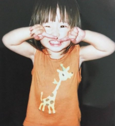 キリンのイラストが描かれたオレンジ色のノースリーブシャツを着て、両手の指を顔の前で立てながら満面の笑みを浮かべる幼少期の菅原咲月。