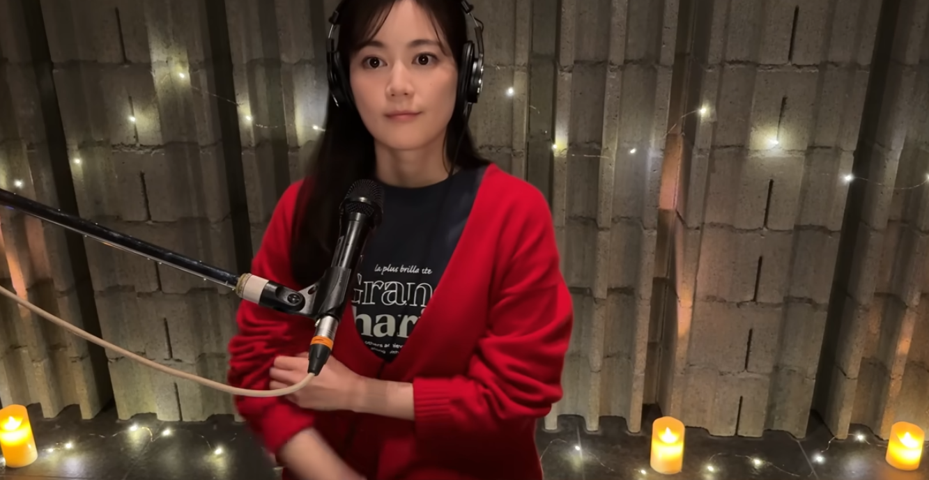 コンクリート壁と温かい電飾が印象的なスタジオで、ヘッドホンを着用し、マイクを前に立つ生田絵梨花の近影。赤いカーディガンを羽織り、真剣な眼差しで音楽と向き合うソロアーティストとしての姿。