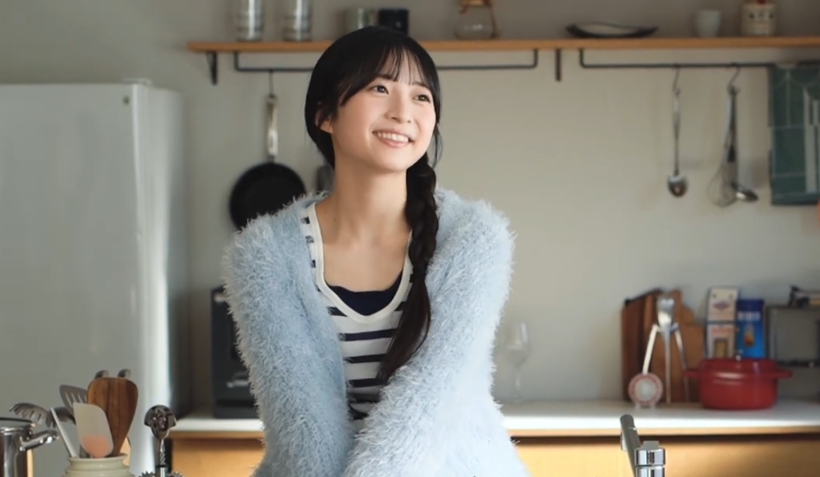 キッチンを背景に、水色のニットカーディガンを羽織って優しく微笑むAKB48 秋山由奈のオフショット風ポートレート。