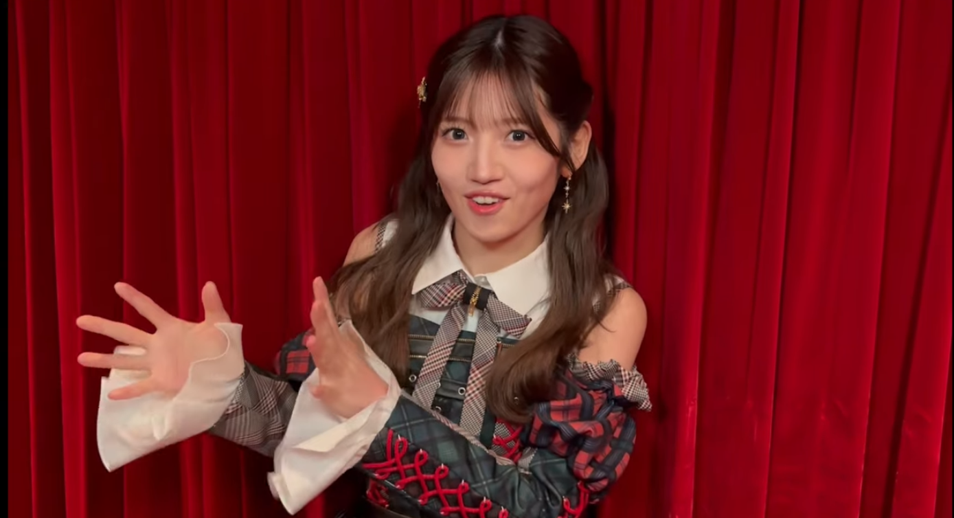 劇場の赤いカーテンを背景に、AKB48のチェック柄の衣装を着て、身振り手振りを交えながら笑顔で話す村山彩希さんの写真