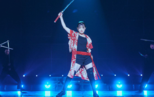 青いステージ照明の中、赤い法被（はっぴ）風の衣装を羽織り、右手で刀を高く掲げて堂々としたポーズをとる村山彩希さんの写真