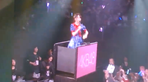 <img src="スクリーンショット 2026-04-19 202027.jpg" alt="AKB48のコンサート会場で、青とピンクのフリルが付いた華やかなアイドル衣装を着用し、ゴンドラに乗ってマイクを持ち、笑顔で歌を歌っている倉野尾成美。背景には、ペンライトを振る多くの観客、ステージ、照明設備が見える。">