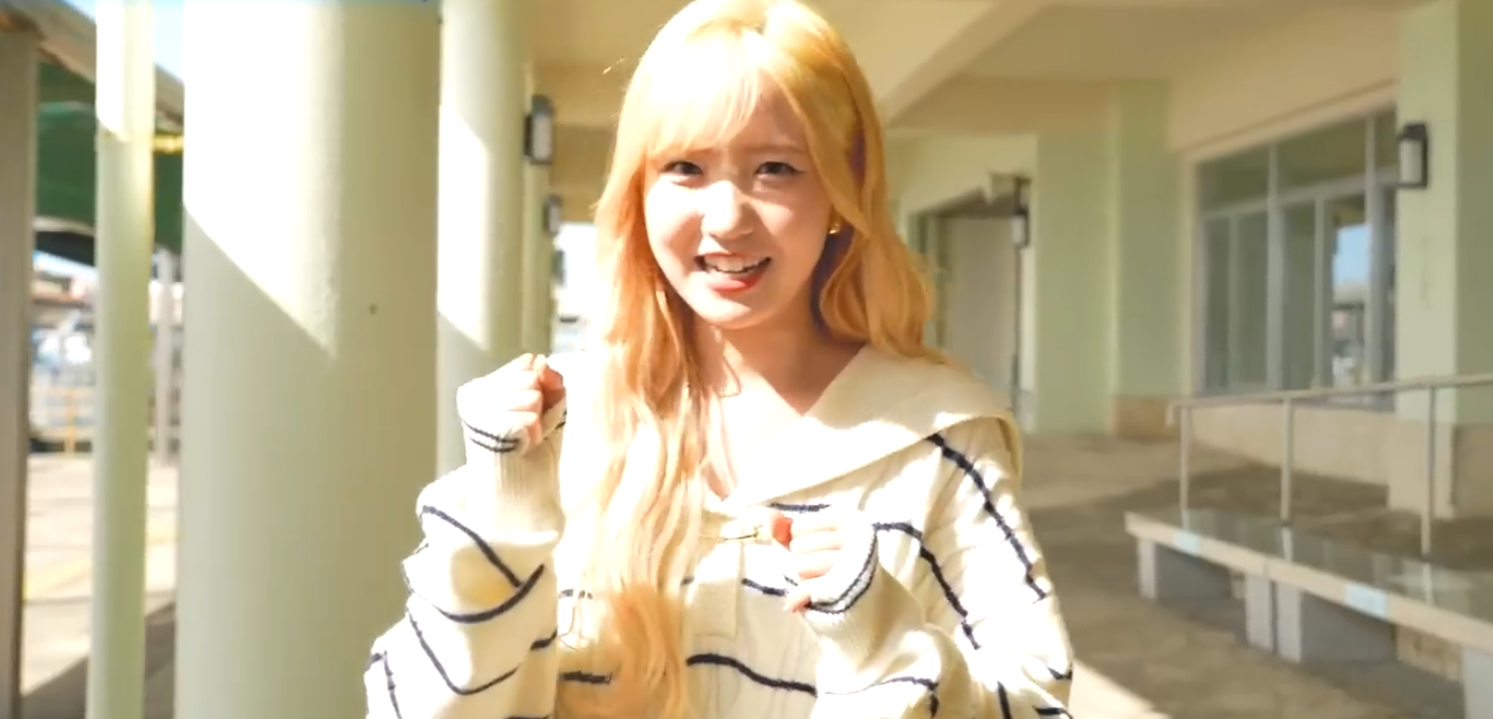 金髪に染め、洗練されたビジュアルに変化したIZ*ONE活動時代の本田仁美。白を基調とした華やかな衣装を身にまとい、ステージ袖で出番を待つ一瞬の微笑みをとらえた写真。胸元にはハングルと日本語で「本田仁美」と書かれた名札を着用しており、世界を舞台に戦う彼女の自信と充実感が伝わる一枚