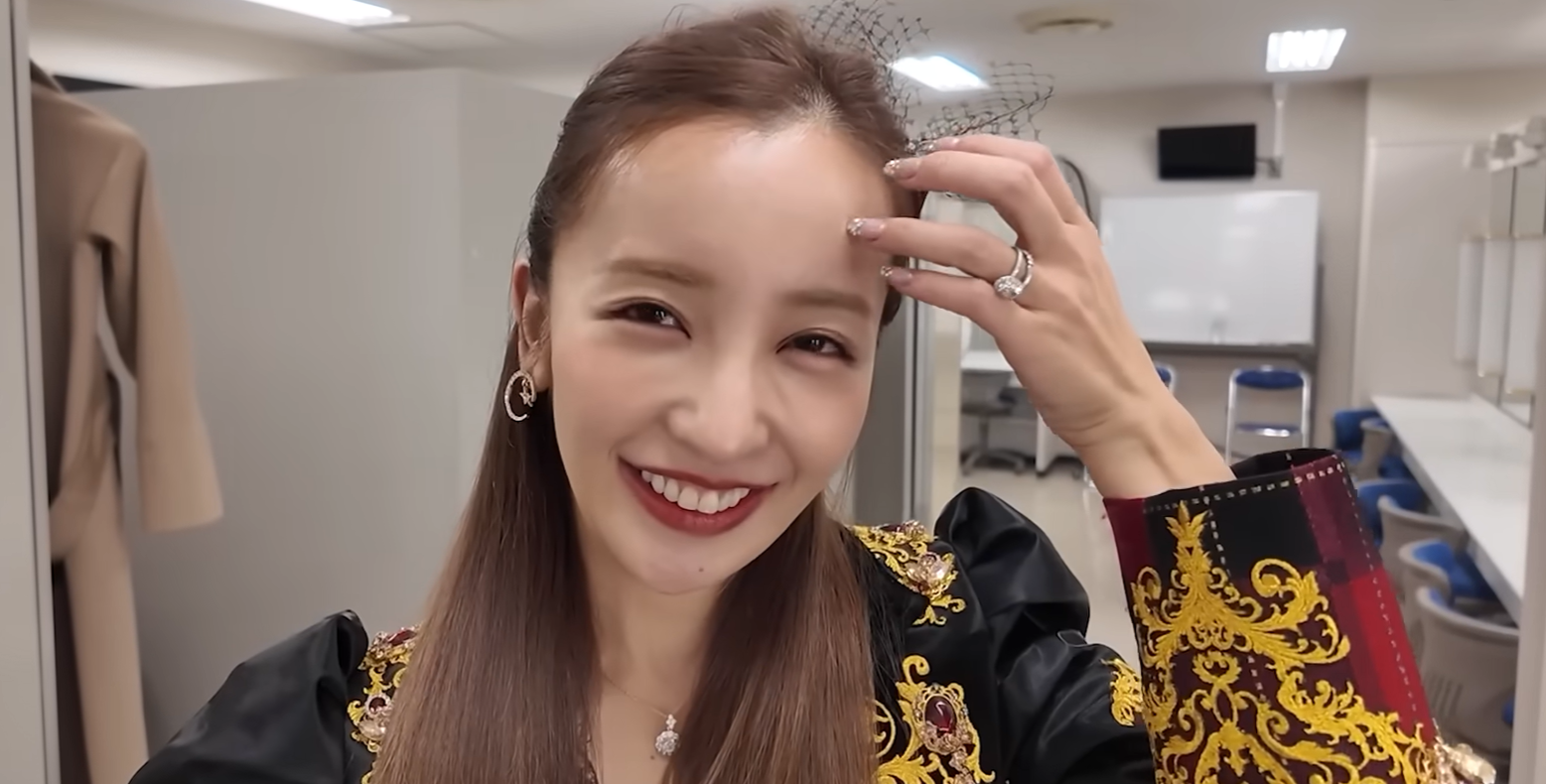 楽屋のような場所で、黒とゴールドの衣装を着て頭の飾りを触りながら笑顔を見せる板野友美