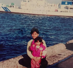 海上保安庁の巡視船を背景に、海辺の堤防で若き日の父に抱きかかえられながらピースサインをする幼少期の篠田麻里子。