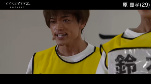 timelesz projectの映像で、黄色いビブスを着て汗を流しながら、歯を見せて充実したような笑顔を浮かべる原 嘉孝。画面右上には「原 嘉孝(29)」のテロップが表示されており、右隣には「鈴」という文字が書かれた名札をつけた別の参加者が一部写り込んでいます。