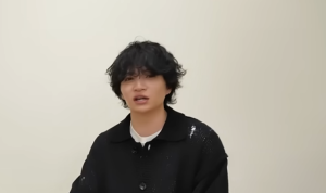 白い背景の前で、黒いニットシャツを着て話す黒髪パーマの菊池風磨。