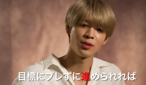 timelesz projectのインタビュー映像で、白いシャツを着て話す金髪の男性。画面下部に「目標にブレずに進めれば」のテロップがある