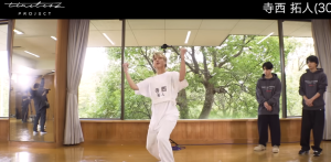 大きな窓から木々が見えるダンススタジオで、「寺西拓人」と書かれた白いTシャツを着てダンスの審査を受ける男性。右奥には見学している他の参加者の姿が写っている。