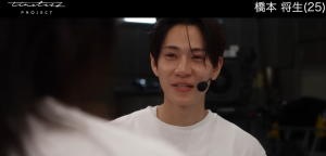 timelesz projectの映像で、白いTシャツにヘッドセットマイクをつけ、涙ぐみながらも少し微笑むような表情で誰かを見つめる男性。画面右上には「橋本 将生(25)」というテロップが表示されています。