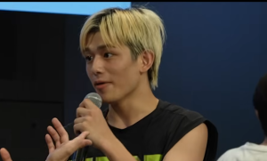 マイクを手に持ち、真剣な表情で話をする金髪(根本が黒色)の男性(篠塚大輝)。左下には、ジェスチャーを交える別の人物の手が一部写り込んでいます。