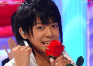 デビュー初期の白い衣装をまとい、手元に赤い一輪の薔薇の花を添えてカメラに弾けるような笑顔を向ける少年時代の松島聡さん