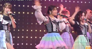 コンサートステージでマイクを持ち、笑顔で手を振りながら歌唱する渋谷凪咲と他のNMB48メンバー