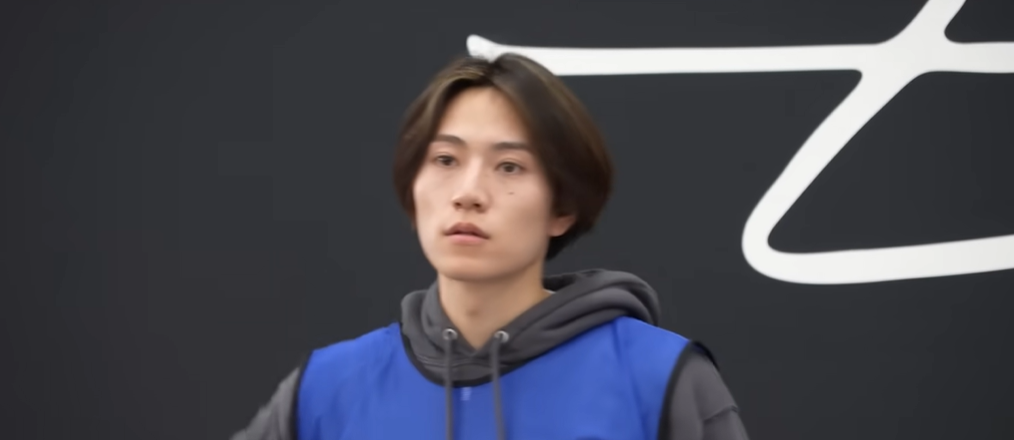 練習中の橋本将生のクローズアップ。橋本将生がグレーのパーカーの上に、大きな「Z」のような白いロゴがプリントされた青いビブスを着用している。彼は前を見つめて立っている。