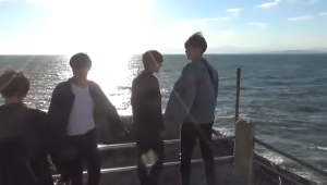 太陽の光が眩しく反射する海を背景に、海沿いのデッキで景色を眺めている3人の若い男性の様子。