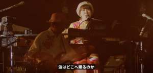 ステージ上でキーボードを弾きながら歌う原由子と、隣でギターを演奏する男性。「波はどこへ帰るのか」の歌詞テロップ