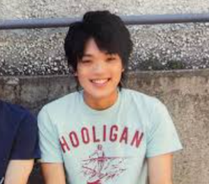 「HOOLIGAN」と書かれた水色のTシャツを着て、屋外で爽快な笑顔を見せる少し長髪の原嘉孝