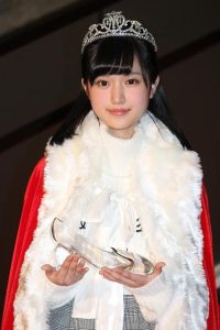 ティアラと赤いマント、白いファーを身につけ、ガラスの靴を持って微笑む福本莉子
