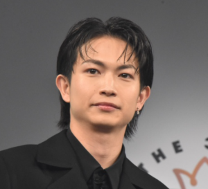 黒のスーツ姿で公の場に登壇し、凛々しい表情でまっすぐ前を見据える綱啓永のバストアップ写真